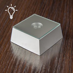 Zdjęcie srebrnej trapezowej podstawki LED. Podstawka ma wysokość trzech centymetrów. U góry zdjęcia, z lewej strony widnieje biała ikonka w kształcie żarówki.