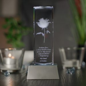 Widok od frontu na podświetloną na biało szklaną statuetkę ustawioną na srebrnej trapezowej podstawce LED. Podstawka ma wysokość trzech centymetrów. W środku statuetki wygrawerowany jest model róży oraz dedykacja z życzeniami na urodziny dla żony. Podstawka stoi na blacie w kolorze ciemnobrązowym. Na drugim planie po obu stronach statuetki widnieją świece.