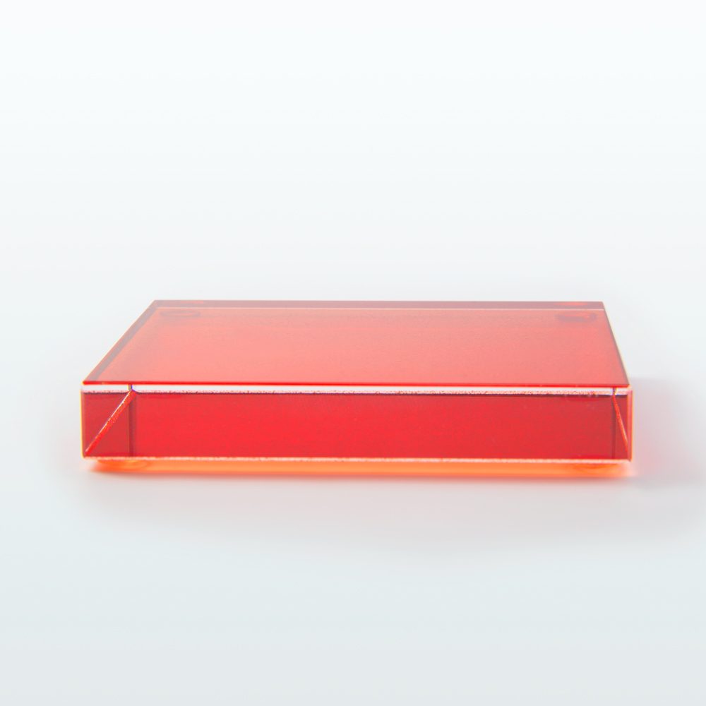 Zdjęcie przedstawiające szklaną podstawkę, zabarwioną na czerwono. Podstawka jest zwrócona frontem do obiektywu. Jej krawędzie są proste, delikatnie zeszlifowane. Podstawka ma wysokość jednego centymetra.