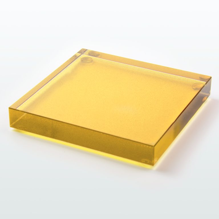 Zdjęcie przedstawiające od góry szklaną podstawkę, zabarwioną na żółto. Podstawka jest delikatnie obrócona frontem od obiektywu. Jej krawędzie są proste, delikatnie zeszlifowane. Podstawka ma wysokość jednego centymetra.