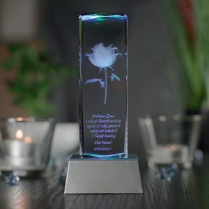 Widok od frontu na podświetloną szklaną statuetkę ustawioną na srebrnej trapezowej podstawce LED. Podstawka ma wysokość trzech centymetrów oraz wielokolorowe światła. W środku statuetki wygrawerowany jest model róży oraz dedykacja z życzeniami na urodziny dla żony. Podstawka stoi na blacie w kolorze ciemnobrązowym. Na drugim planie po obu stronach statuetki widnieją szklane świeczniki z palącymi się podgrzewaczami.