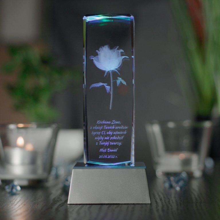 Widok od frontu na podświetloną szklaną statuetkę ustawioną na srebrnej trapezowej podstawce LED. Podstawka ma wysokość trzech centymetrów oraz wielokolorowe światła. W środku statuetki wygrawerowany jest model róży oraz dedykacja z życzeniami na urodziny dla żony. Podstawka stoi na blacie w kolorze ciemnobrązowym. Na drugim planie po obu stronach statuetki widnieją szklane świeczniki z palącymi się podgrzewaczami.