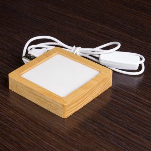 Zdjęcie drewnianej podstawki LED z mleczką nakładką. Podstawka ma wysokość dwóch centymetrów. Za podstawką leży kabel USB dedykowany do niej.